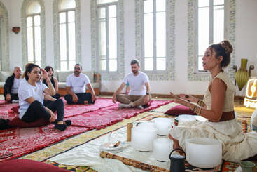 A woman leads a sound healing session with singing bowls, while a group of people sit on mats and cushions, listening attentively in a bright, serene room.