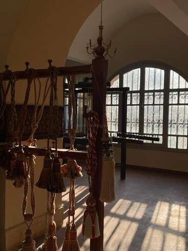 Sunlight softly illuminates an elegant room with arched windows, where decorative tassels hang from a wooden rack, casting intricate shadows across the tiled floor. Peaceful ambiance.