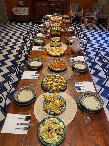 A long wooden table is set for a meal with colorful dishes, salads, eggs, pastries, and ceramic bowls, surrounded by chairs and patterned tile flooring.