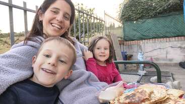 A smiling woman and two happy children sit outdoors, enjoying a meal with tortillas or pancakes. The background shows a fence, greenery, and a casual setting.