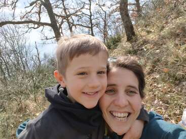 A joyful woman and young boy smile closely together outdoors in a forest setting. Leafless trees and dry grass surround them on a bright, cheerful day.