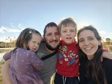 A happy family of four poses outdoors, smiling at the camera. The sun shines behind them. The little girl appears shy, while the boy looks cheerful.