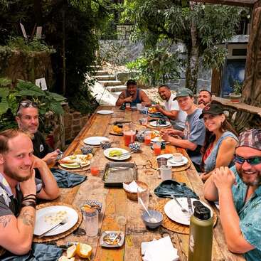 A group of friends enjoys a meal outdoors at a rustic wooden table surrounded by greenery, smiling and sharing food, drinks, and friendly conversation in natural light.