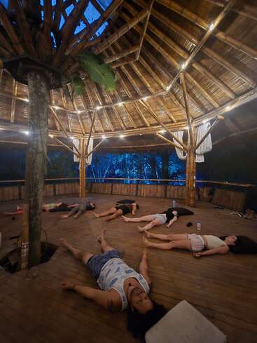 Under a bamboo pavilion at dusk, several people practice savasana, lying on wooden floor mats, surrounded by nature, exuding peace and tranquility in the ambiance.