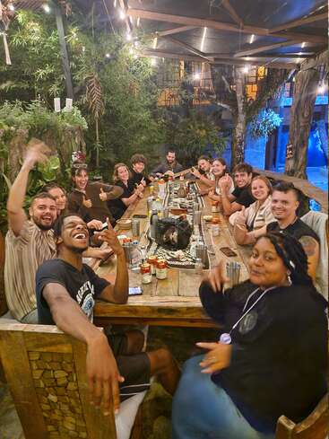 Un grupo de amigos diversos se sientan alrededor de una mesa rústica de madera al aire libre, riendo y sonriendo, disfrutando de bebidas y comida bajo luces parpadeantes en un entorno acogedor y exuberante.