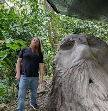 Un hombre con una larga barba pelirroja se encuentra en un frondoso bosque verde junto a un gran rostro esculpido que se asemeja a un anciano barbudo, hecho de arcilla.