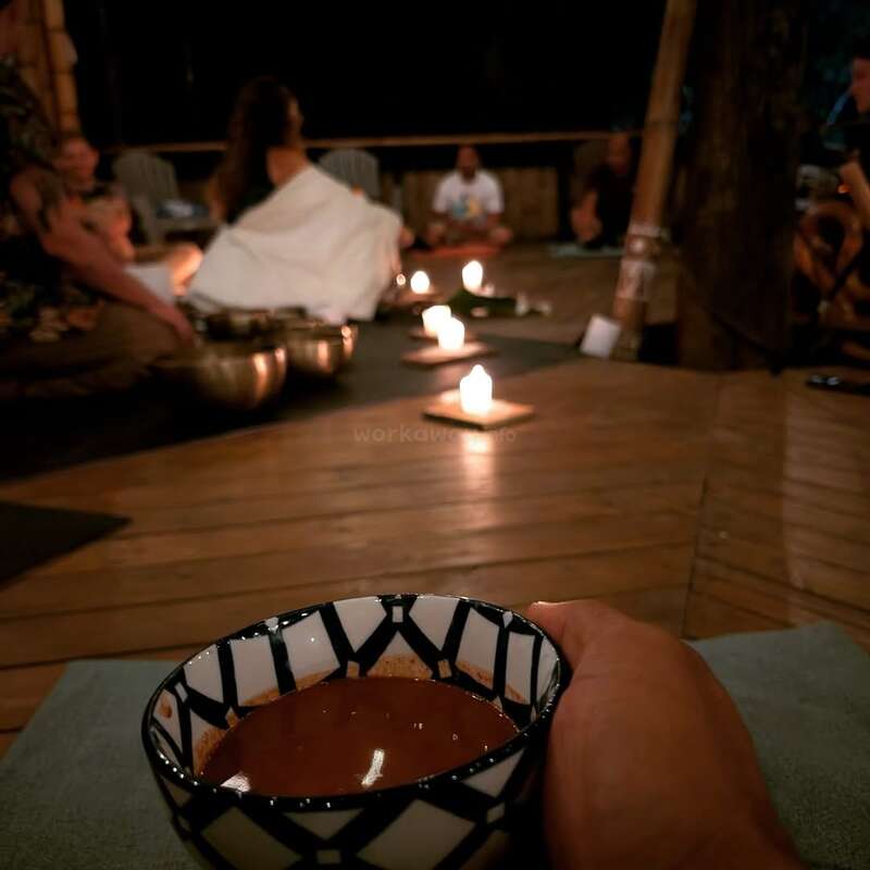 A person holds a patterned bowl with a dark liquid, sitting in a circle around lit candles. Others are gathered on mats, creating a serene atmosphere.