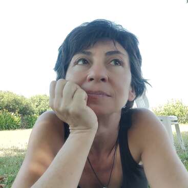 Una mujer de pelo corto y oscuro está tumbada sobre la hierba, apoya la barbilla en la mano y mira pensativa a lo lejos. Está rodeada de vegetación y una silla.