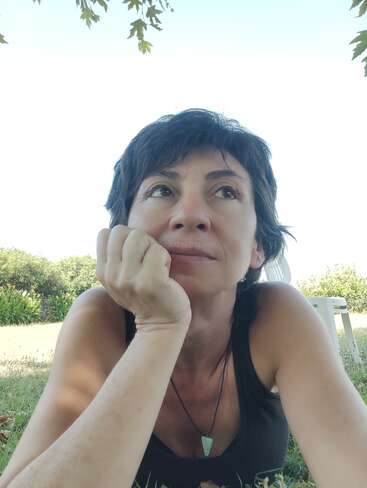 A woman with short dark hair lies on grass, resting her chin on her hand, gazing thoughtfully into the distance. Greenery and a chair surround her.