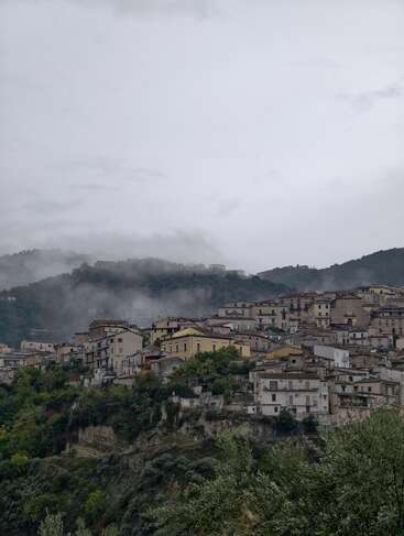 Un brumoso pueblo de montaña descansa tranquilamente bajo un cielo nublado. Las casas de piedra se agrupan en una ladera verde, rodeadas de frondosos árboles y envueltas en la niebla matinal.