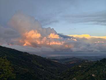 Unas montañas verdes y ondulantes enmarcan un vasto valle, mientras unas nubes espectaculares, iluminadas con tonos naranjas y rosas por el sol poniente, dominan el cielo. Una vista apacible y sobrecogedora.