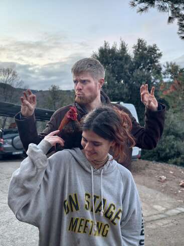 A young woman in a grey hoodie with “ON STAGE MEETING” stands outside, smiling. A man behind her poses playfully while a chicken perches on her shoulder.