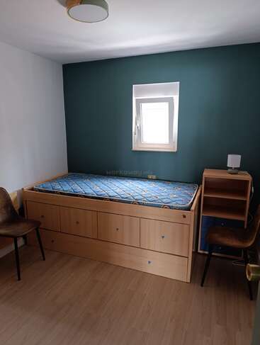 Esta habitación tiene una cama individual con cajones, dos sillas marrones, una mesita de noche de madera con estantes, una lámpara, suelo de madera y una pequeña ventana.