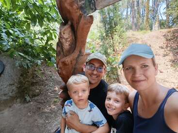 Una sonriente familia de cuatro miembros posa al aire libre delante de una escultura de madera con forma de árbol en la que aparece un búho. La luz del sol se filtra a través de los árboles, creando un ambiente alegre.