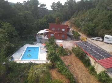 En esta imagen se ve una casa roja rodeada de árboles, una piscina con gente, paneles solares, un coche aparcado, una autocaravana y un camino de tierra sinuoso.