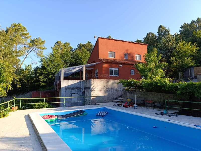 Una casa roja de dos pisos se alza detrás de una piscina azul cristalina rodeada de vegetación y árboles. En el agua flotan juguetes para la piscina. Es una tarde tranquila y soleada.