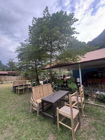Área de estar ao ar livre com móveis de bambu, cercada por grama verde e árvores, sob um céu nublado. Ambiente aconchegante e rústico ao lado de um prédio com telhado de zinco.