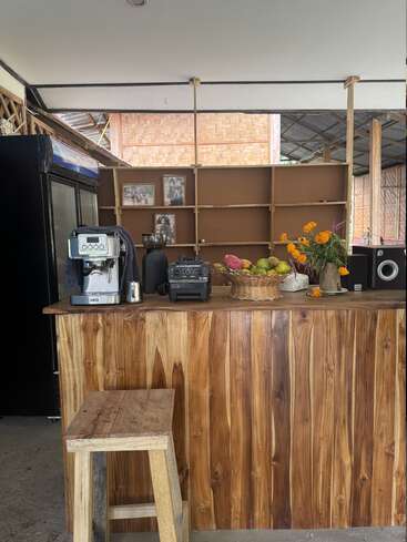 Esta imagem mostra um balcão rústico de madeira com um banquinho, uma máquina de café, um liquidificador, uma cesta de frutas, flores, prateleiras vazias, fotos emolduradas e uma geladeira. Ambiente aconchegante.