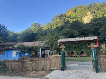 Der rustikale Eingang einer Herberge mit Bambuszaun, Holzschildern und kleinen dreieckigen Bungalows. Umgeben von üppig grünen Bäumen und Hügeln unter einem strahlend blauen Himmel.