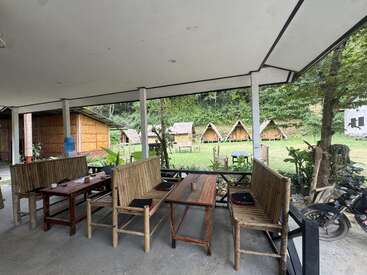 Cette photo montre un coin salon en plein air avec des meubles en bambou, donnant sur une pelouse et de petites cabanes en bois, le tout entouré d'une végétation luxuriante et d'arbres. L'atmosphère y est paisible.