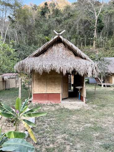Eine kleine Bambushütte mit Strohdach steht auf einem grasbewachsenen Hof. In der Nähe wachsen Bananenstauden, und im Hintergrund erheben sich bewaldete Hügel. Eine friedliche Szene.