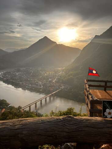 Una impresionante puesta de sol ilumina un valle con un río, un puente, las montañas al fondo, un pueblo a sus pies y una bandera de Laos ondeando en un mirador de madera que domina la escena.