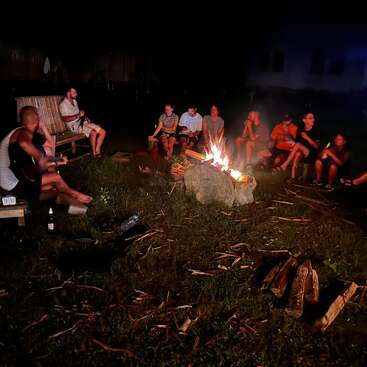 Un grupo de personas se sienta alrededor de una hoguera que arde en la noche, charlando, riendo y tocando la guitarra, disfrutando de una acogedora velada al aire libre bajo el cielo oscuro.