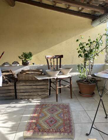 Sonnige Terrasse mit einem rustikalen Stuhl, zwei Schüsseln auf einem Tisch, Holzkisten, Topfpflanzen, einem geflochtenen Korb und einem bunten Teppich mit Muster auf dem Fliesenboden.
