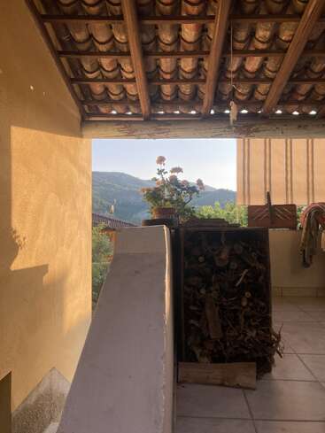 Ein gemütlicher Balkon mit einem Ziegeldach, Topfpflanzen, gestapeltem Brennholz und malerischen Bergen im Hintergrund, der in sanftes, warmes Sonnenlicht getaucht ist und eine ruhige Atmosphäre schafft.