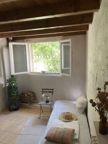 Dieses gemütliche Zimmer hat eine Holzdecke, ein offenes Fenster mit Blick auf den Garten, ein Tagesbett, einen rustikalen kleinen Tisch, einen Hut, eine Holzbox, Pflanzen und getrocknete Blumen in einer Vase.
