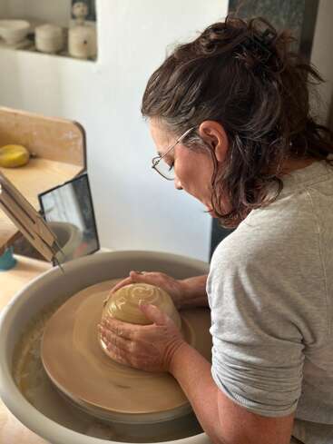 A woman with glasses and curly hair carefully shapes a bowl on a pottery wheel, focused and creative, in a bright, cozy ceramic studio space.