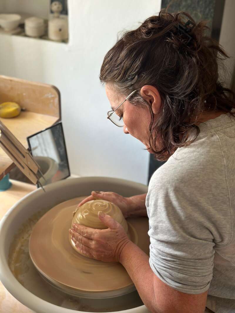 Eine Frau mit Brille und lockigem Haar formt sorgfältig eine Schale auf einer Töpferscheibe, konzentriert und kreativ, in einem hellen, gemütlichen Keramikstudio.