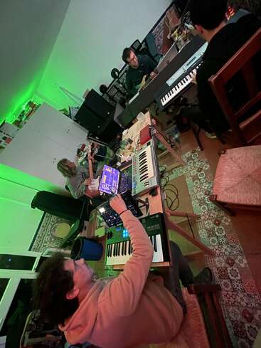 Quatro pessoas estão em uma sala aconchegante com iluminação verde, tocando sintetizadores, teclados e um violão, cercadas por equipamentos de música, laptops e piso de azulejos estampados.