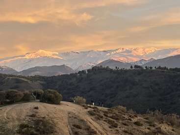 Montanhas cobertas de neve brilhando sob um pôr do sol dourado, com colinas verdes ondulantes abaixo. As pessoas caminham por uma trilha de terra, curtindo a vista serena e pitoresca da paisagem.