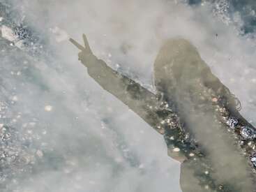 Das Bild zeigt die Silhouette eines Menschen, der seine Arme zum Friedenszeichen erhebt und sich in einer Pfütze mit gräulichem Wasser und verstreuten Trümmern spiegelt.
