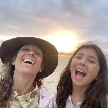 Das Bild zeigt zwei Frauen, die bei Sonnenuntergang am Strand lachen, während die Sonne hinter ihnen untergeht und der Himmel bewölkt ist.