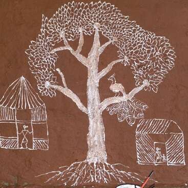 This image shows traditional Warli art, featuring a big tree, a peacock, two huts with human figures, all painted in white on a brown wall.
