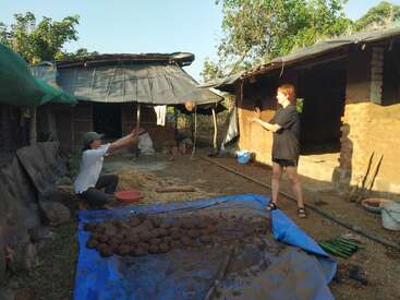 La imagen muestra a dos individuos en el exterior de un edificio rústico, con una lona azul cubierta de barro y bolas de arcilla en primer plano.