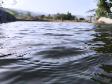 Uma imagem em close-up de águas calmas e ondulantes, talvez um lago ou lagoa, com árvores, colinas e o céu desfocados ao fundo, criando uma cena tranquila.