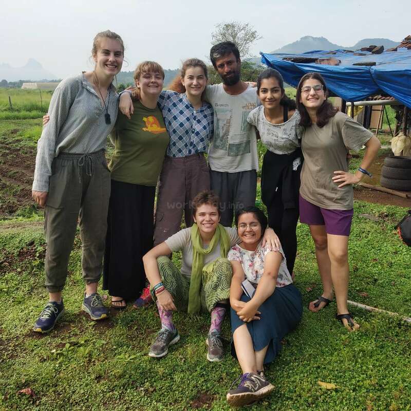 The image depicts a group of eight people posing for a photo on a grassy field, with a blue tarp-covered structure and a rural landscape in the background.