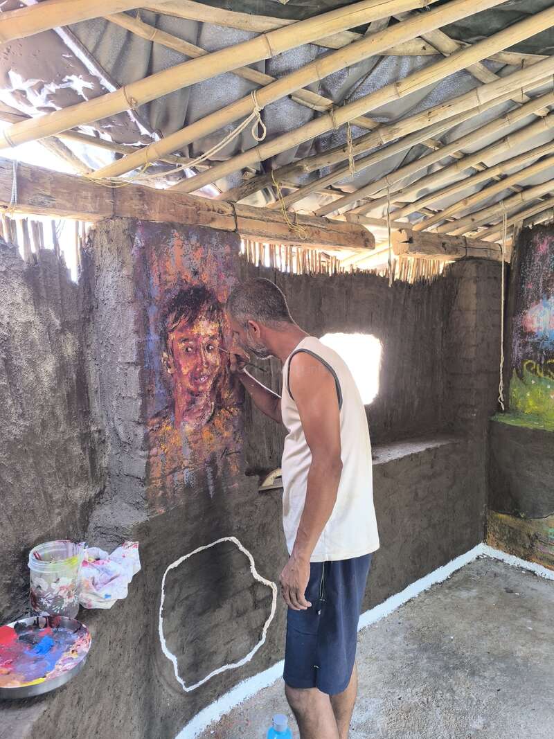 Um homem vestindo uma camiseta sem mangas e shorts pinta um retrato colorido em uma parede rústica dentro de um prédio com estrutura de bambu, com vigas à mostra e luz do sol entrando.