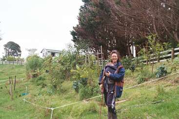 Una mujer sonriente está de pie en una ladera cubierta de hierba con un bebé en un portabebés delantero. Detrás de ella se ven plantas frondosas, árboles y una valla blanca.