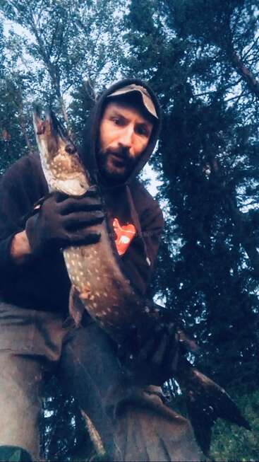 A man in outdoor clothing and gloves proudly holds a large fish, standing in a forested area with tall trees and a dusky sky behind him.