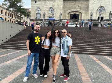 L'image représente quatre personnes debout devant un grand bâtiment en pierre avec des escaliers, posant pour une photo, entourées d'un paysage urbain avec des personnes et des bâtiments à l'arrière-plan.