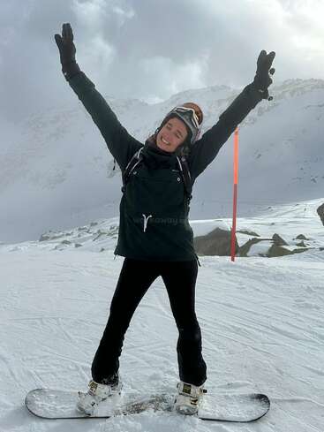 Une femme se tient debout sur un snowboard, les bras levés, portant un casque, des gants et des vêtements d'hiver, sur fond de montagne enneigée.
