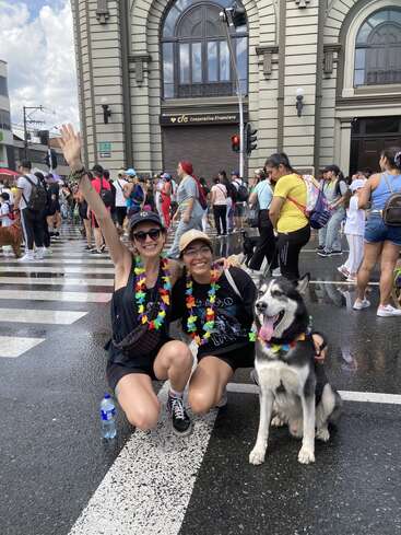 Deux femmes et un chien posent dans une rue de la ville, parés de leis colorés, devant une foule et un grand bâtiment sur lequel on peut lire "CFA Cooperativa Financiera".