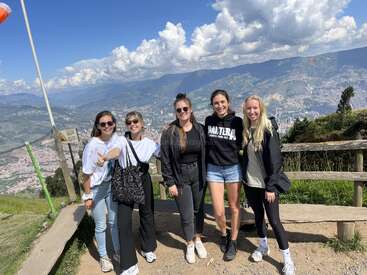 L'image représente cinq femmes posant pour une photo au sommet d'une montagne, avec un paysage urbain et des montagnes visibles à l'arrière-plan sous un ciel bleu avec des nuages.