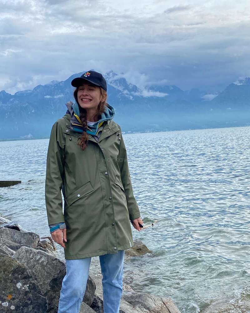 L'image représente une femme debout sur des rochers au bord d'un lac, vêtue d'une veste verte, d'un jean bleu et d'une casquette noire, avec des montagnes visibles à l'arrière-plan sous un ciel nuageux.