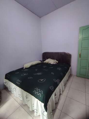 This image shows a small, simple bedroom with a double bed, dark headboard, two pillows, green bedspread, white skirt, tiled floor, and green door.
