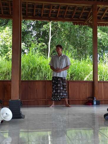 A person stands barefoot on a tiled floor in a wooden pavilion, wearing a patterned sarong and light shirt, surrounded by lush green outdoor vegetation.